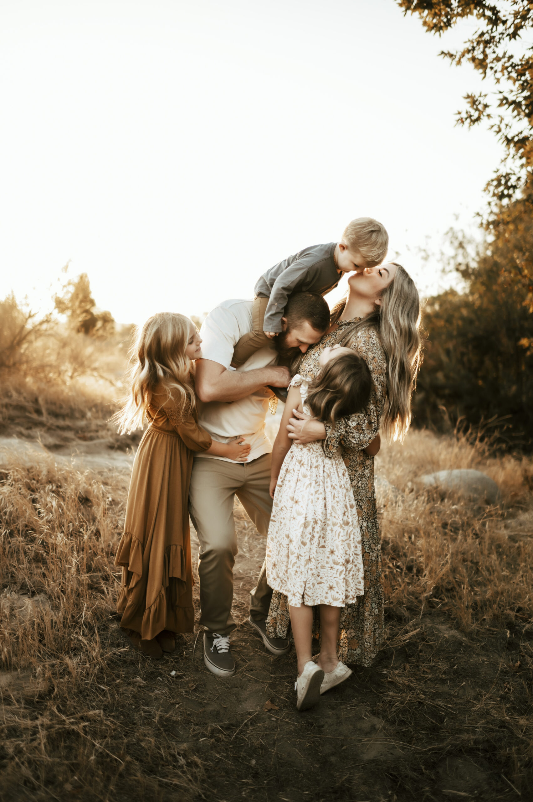 mom kissing baby that’s on dad’s shoulders while daughters are hugging mom and dad in an autumn setting