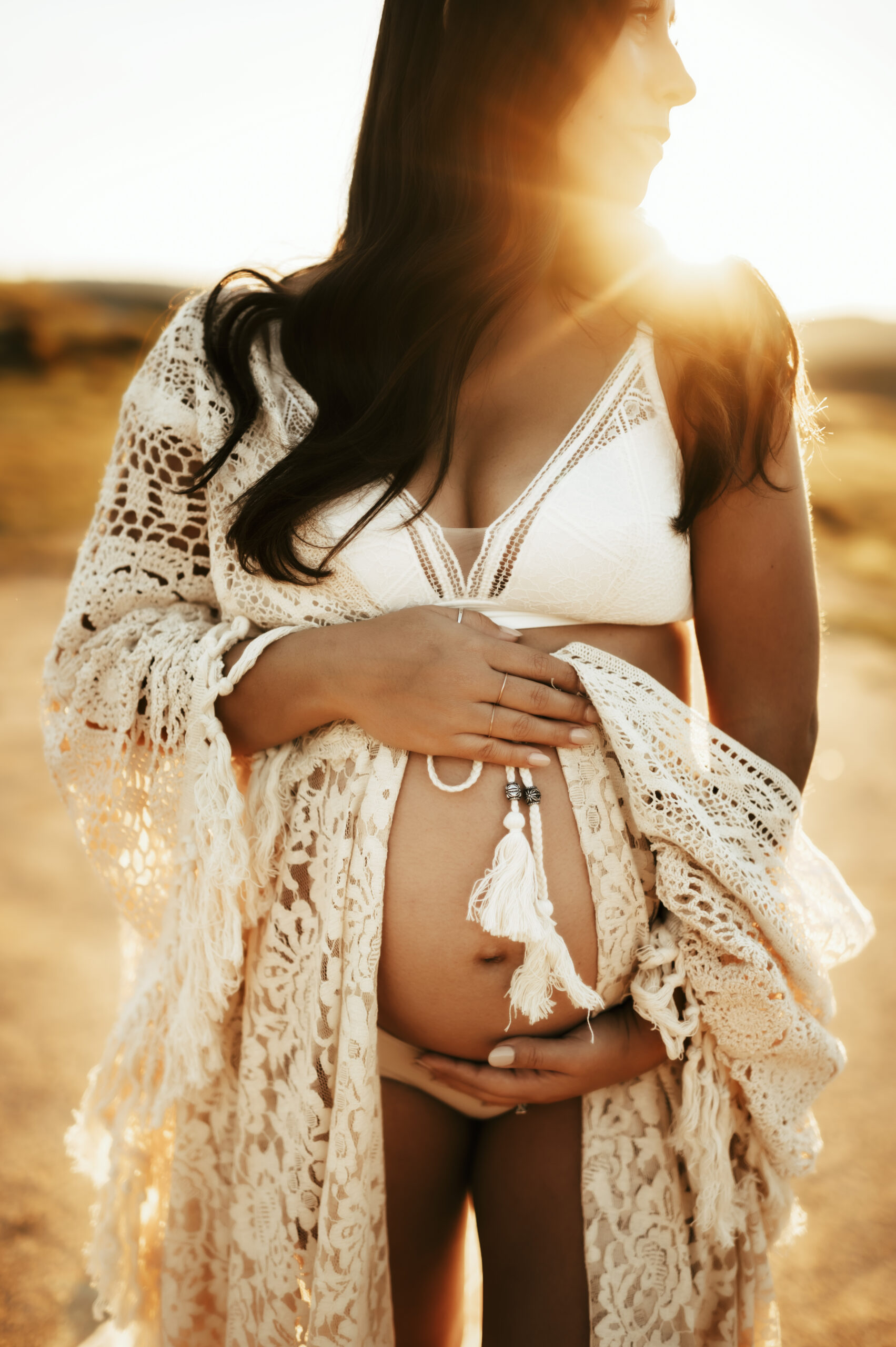 Pregnant mother looking off in the distance, holding her baby in a crocheted lace robe with sun rays behind her